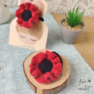 May include: Two red crocheted poppy flowers with black centers. The flowers are attached to hair elastics. The flowers are on a wooden surface.