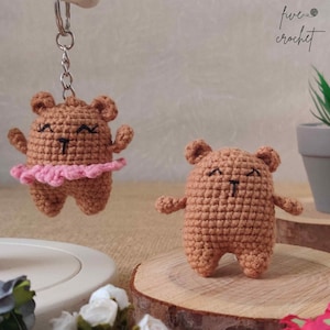 May include: Two crocheted brown bear keychains. One bear is wearing a pink tutu and is attached to a silver keychain. The other bear is standing on a wooden surface.