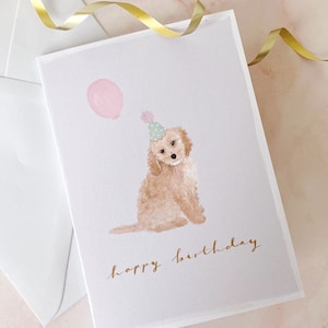 May include: A birthday card featuring a watercolour illustration of a dog wearing a party hat and holding a pink balloon. The card reads "happy birthday" in gold script.