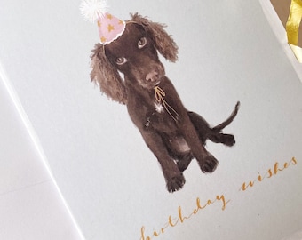May include: A birthday card featuring a brown dog wearing a pink and gold party hat. The card has the words "birthday wishes" written in gold lettering.