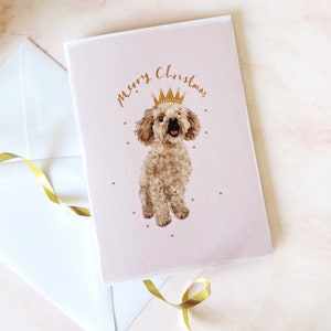 May include: A pink Christmas card with a white border featuring a brown poodle wearing a gold crown. The card says "Merry Christmas" in gold lettering.
