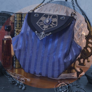 May include: A blue and silver striped fabric purse with a black leather flap and silver metal embellishments. The purse has a chain strap and is decorated with a silver metal design.