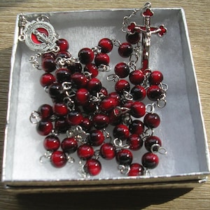 May include: A rosary featuring round, polished beads with a deep red and black gradient pattern, connected by silver-toned metal links. The rosary includes a silver-toned crucifix with red enamel accents and a circular medal with a red center.