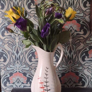 May include: A cream-coloured ceramic pitcher with a curved handle and a flared spout, featuring a stylised black and red botanical branch pattern on the front, filled with a bouquet of yellow and deep purple lisianthus flowers and green foliage.