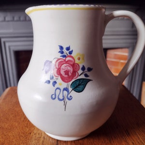 May include: White ceramic jug with a floral design featuring a pink rose, blue and yellow flowers, and green leaves. The jug has a yellow rim and a light blue handle.