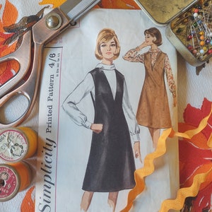 May include: A Simplicity sewing pattern for a dress, featuring a black and white illustration of a woman wearing a black dress with a white collar and white trim. The pattern number is 4/6. The pattern is on a white background with a yellow ribbon.