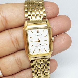 Può includere: Un orologio Orient color oro con quadrante bianco e cinturino in metallo dorato. Il quadrante dell'orologio ha indici orari romani neri, lancette nere e una finestrella per la data alle 3. L'orologio è resistente all'acqua fino a 50 metri.