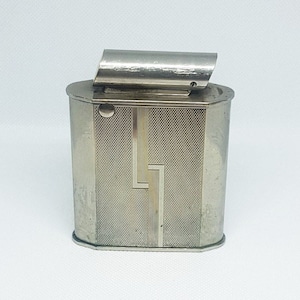 May include: A silver lighter with a geometric design on the front. The lighter has a hinged lid and a rounded shape.