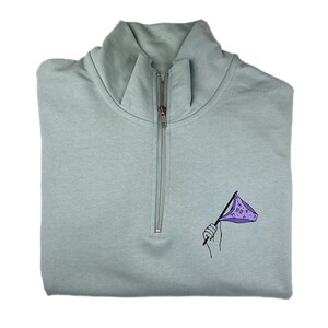 Könnte beinhalten: Ein hellgrüner Viertelreißverschluss-Pullover mit einer Schwarzweißillustration einer Hand, die eine lila Flagge mit dem Text "B.I.G." hält.