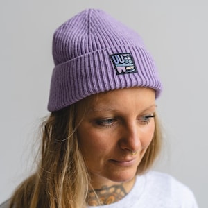 Könnte beinhalten: Eine lila Beanie-Mütze mit gerippter Strickstruktur. Auf der Mütze befindet sich ein rechteckiger Aufnäher mit dem Text "UU" und einem grafischen Design. Die Person, die die Mütze trägt, hat blonde Haare und einen Nasenring.