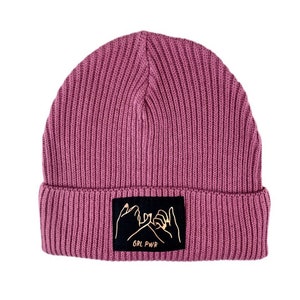 Könnte beinhalten: Rosa gestrickte Mütze mit einem schwarzen Patch, auf dem zwei goldene Hände ein Pinky-Versprechen machen und der Text "GRL PWR" steht.