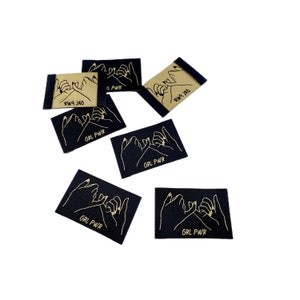 Könnte beinhalten: Sechs schwarze Stoff-Etiketten mit goldener Beschriftung und einer Grafik von zwei Händen, die ein Pinky-Versprechen machen. Die Etiketten lauten "GRL PWR".