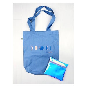 Könnte beinhalten: Eine blaue Tragetasche mit einem Design aus fünf Monden und dem Text "Just a Phase" in Weiß. Vor der Tasche liegt eine kleine, regenbogenfarbene Tasche mit Reißverschluss.