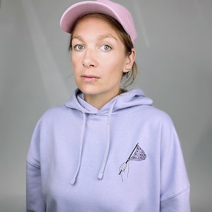 Könnte beinhalten: Eine Person trägt einen lilafarbenen Hoodie mit einer kleinen Grafik einer Hand, die eine lila-weiße Flagge hält. Die Person trägt auch eine rosa Baseballkappe.