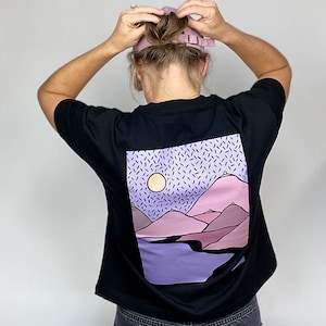 Könnte beinhalten: Schwarzes T-Shirt mit einem grafischen Aufdruck einer lila und pinkfarbenen Berglandschaft mit einer gelben Sonne und schwarzen Streuseln auf einem lila Hintergrund.