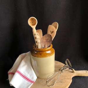 May include: A selection of wooden kitchen utensils, including spoons and spatulas, in a tan and brown ceramic jar. A white and red striped tea towel and a metal whisk sit on a wooden chopping board, against a black backdrop.