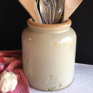 May include: A collection of kitchen utensils in a beige ceramic crock. The utensils include wooden spoons, a metal whisk, and a metal spoon. A red and white checkered cloth and a garlic bulb are in the foreground.