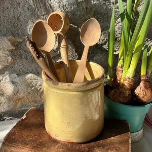 May include: A yellow ceramic crock containing three wooden kitchen utensils, including two spoons and a serrated tool, resting on top of a weathered, leather-bound book. The crock is cylindrical with a smooth, glazed finish and a slightly worn base.