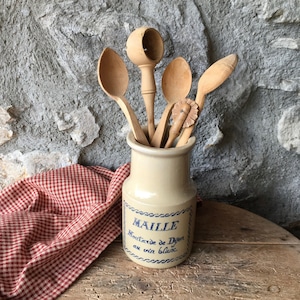 May include: A collection of wooden kitchen utensils, including spoons and a nutcracker, displayed in a beige ceramic jar. The jar has blue text that reads "MAILLE Moutarde de Dijon au vin blanc". A red and white checkered cloth is draped nearby.