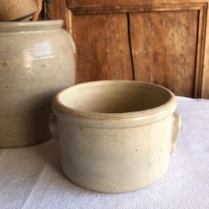 May include: A small, cream-coloured ceramic bowl with two small handles. The bowl is round with a slightly flared rim. A larger, similar-coloured ceramic jar is behind the bowl. The background is a wooden door.