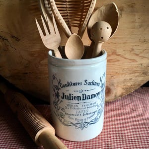 以下が含まれることがあります： フォーク、スプーン、ザルなどの木製キッチン用品のコレクション。青い文字が入った白い陶器の瓶に収められています。木製のめん棒が赤と白のチェック柄の表面に置かれています。瓶には「Confitures Surlines Julien Damoy」と書かれています。