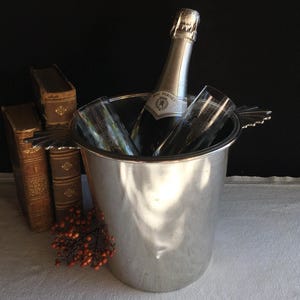 Op de afbeelding: Een zilveren champagne-emmer bevat een fles champagne en twee fluiten. De emmer staat op een wit tafelkleed, met antieke boeken en oranje bessen op de achtergrond. De champagnefles heeft een zilveren etiket.