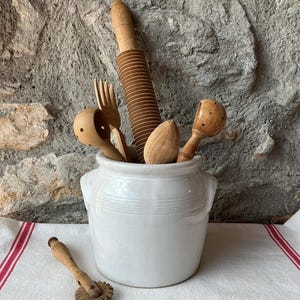 Peut inclure: Un ensemble d'ustensiles de cuisine anciens en bois, dont un rouleau à pâtisserie, une fourchette et une cuillère, sont présentés dans un pot en céramique blanc. Une roulette à pâtisserie repose sur un tissu blanc à rayures rouges. En arrière-plan, un mur en pierre.