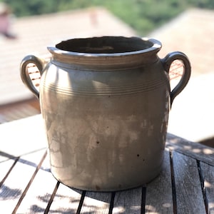 May include: A beige ceramic pot with two side handles and a wide opening. The exterior features a smooth, glazed finish with subtle horizontal ridges near the rim. The vessel has a cylindrical body with a slightly tapered base and visible surface texture.