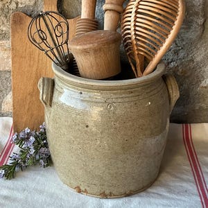 May include: A beige stoneware crock with two side handles containing a collection of kitchen tools including a wire whisk, a wooden masher, a wooden rolling pin, and a woven rattan whisk, with a wooden cutting board positioned behind the crock.