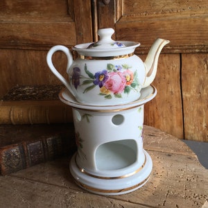 May include: A white porcelain teapot with a floral design, sitting on a matching warmer. The teapot has a lid and spout with gold trim. The warmer has a circular opening and gold accents. The floral pattern includes pink, purple, and yellow flowers.