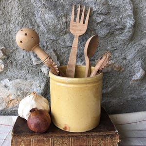 Pode incluir: Um pote de cerâmica amarelo contém utensílios de cozinha de madeira, incluindo um garfo, uma colher e uma ferramenta ranhurada. Um bulbo de alho e um pequeno objeto redondo marrom-avermelhado repousam sobre um livro antigo.