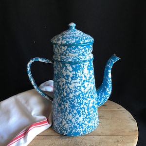 Puede incluir: Una cafetera vintage de esmalte azul y blanco moteado con tapa y asa. La cafetera está sobre una superficie de madera con una toalla blanca y roja.