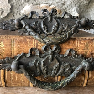 May include: Two antique bronze drawer pulls with ornate floral and ribbon designs. The pulls are set against a backdrop of aged, leather-bound books, suggesting a vintage or historical theme. The metal has a weathered appearance.