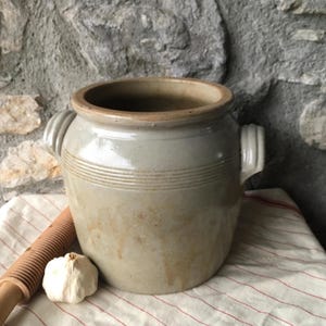 May include: A vintage, off-white stoneware crock with two side handles and horizontal banded detailing. The antique jar is displayed with a garlic bulb and a wooden kitchen tool on a red and white striped cloth.