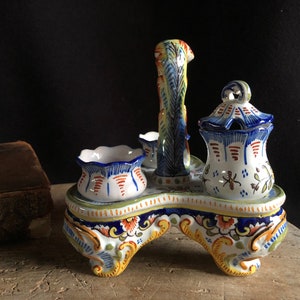 May include: A decorative ceramic condiment set with a handle, featuring a white base with blue, orange, and yellow floral patterns. The set includes a lidded jar, two small bowls, and a central handle, all with hand-painted details.
