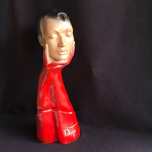 Puede incluir: Una cabeza de maniquí vintage con los ojos cerrados, sostenida por un brazo y una mano rojos. La cabeza está pintada con un tono de piel neutro, cabello negro, y el brazo rojo tiene el texto "Christian Dior" en letras doradas. El maniquí es un artículo decorativo.