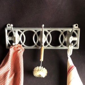 Op de afbeelding: Een zilverkleurige metalen wandkapstok met drie haken en een decoratief geometrisch ontwerp. Een rood-wit geruite doek, een flessenborstel en een wit-rood gestreepte doek hangen aan de haken.