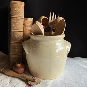May include: A cream-coloured ceramic utensil pot with handles, holding wooden spoons and a fork. Next to it are antique books with gold lettering and a wooden pastry cutter. A small pomegranate sits on the table.