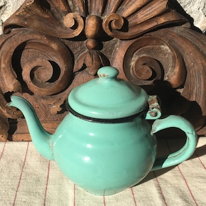 May include: A turquoise enamel teapot with a lid and a curved spout. The teapot has a black trim and a rounded handle. The background features a carved wooden decorative element.