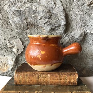 Op de afbeelding: Een kleine, ronde, bruine keramische pot met een handvat en een lichtgekleurde rand. De pot staat op twee antieke boeken met versleten, getextureerde covers. De achtergrond is een ruwe, grijze stenen muur.