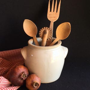 Puede incluir: Una vasija de cerámica color crema que contiene dos cucharas de madera, un tenedor de madera y una rueda de repostería de madera. Dos granadas secas descansan frente a la vasija, parcialmente cubierta por una servilleta de cuadros rojos y blancos.