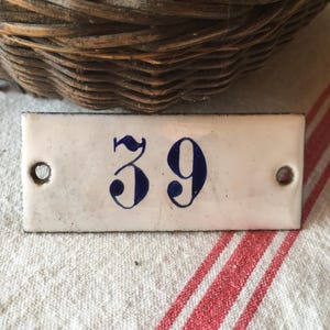 Könnte beinhalten: Rechteckiges weißes Emaille-Schild mit der Zahl 39 in Dunkelblau. Das Schild hat zwei Löcher zur Befestigung und einen schwarzen Rand. Der Hintergrund umfasst einen Weidenkorb und einen rot-weiß gestreiften Stoff.