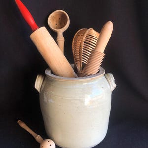May include: A collection of vintage kitchen utensils, including a rolling pin with a red handle, a wooden spoon, and a whisk, are displayed in a light green ceramic crock. A wooden muddler rests in front of the crock. The background is black.