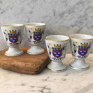 May include: Four white porcelain egg cups with gold rims and decorative crests. Each cup features a detailed emblem with a crown, figures, and a shield with animals. The cups are displayed on a wooden board.