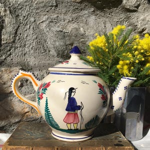 May include: Cream-colored ceramic teapot with a hand-painted design. The teapot features a figure in traditional clothing, floral accents, and a blue lid. The handle is orange and the spout is white. The teapot is sitting on a vintage book.