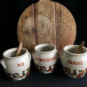 May include: Three white ceramic jars with handles, labeled "RIZ", "CORNICHONS", and "FARINE" in red lettering. Each jar features a rooster and floral design. Wooden spoons rest inside the jars, with a wooden cutting board in the background.