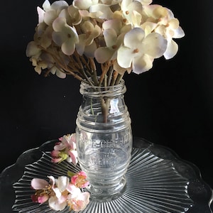 Pode incluir: Uma garrafa de vidro transparente com um buquê de flores brancas e roxas claras. A garrafa está em um prato de vidro transparente e canelado, com pequenas flores rosa e brancas. A garrafa tem texto em relevo. Um arranjo floral vintage.