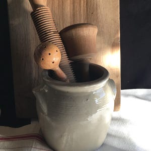 Op de afbeelding: Een verzameling vintage keukengereedschappen, waaronder een houten citroenpers, een geribbeld houten gereedschap en een houten stamper, zijn gerangschikt in een grijze keramische pot. Een houten snijplank staat op de achtergrond.