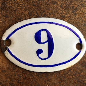 Peut inclure: Plaque ovale en émail blanc avec le chiffre neuf bleu et un liseré bleu. La plaque comporte deux trous de fixation. Le fond est une surface marron texturée.