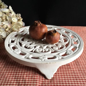 Puede incluir: Un salvamanteles blanco de hierro fundido con un diseño calado ornamentado, que sostiene dos granadas marrones. El salvamanteles tiene un asa decorativa y está sobre un mantel a cuadros rojos y blancos. Se ven acentos florales decorativos.